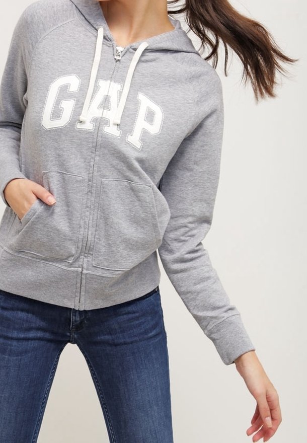 Sudadera gris con cremallera y el logo blanco de "GAP", capucha con cordón, bolsillos frontales y puños acanalados. Combinada con jeans azules.
