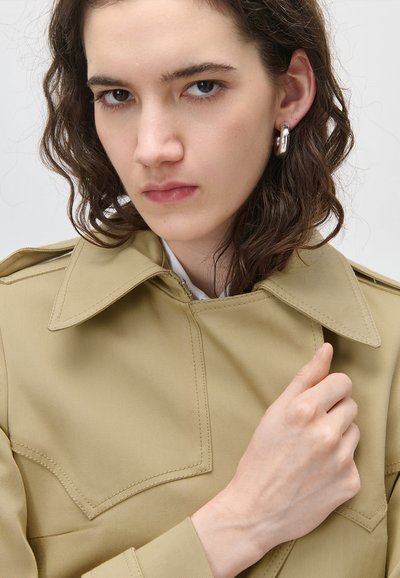 Νεαρός με κυματιστά καστανά μαλλιά που φοράει μπεζ παλτό και ασημένιο σκουλαρίκι σε σχήμα κρίκου, κρατώντας το γιακά του παλτού με το χέρι μπροστά σε φωτεινό φόντο.