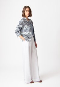 Gaiši zila un balta jaka ar sarežģītu rakstu, kombinēta ar baltām platajām lina biksēm un brūnām sandālēm, stāvoša uz vienkārša fona.