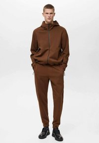 Smeđi zip-up dukserica i odgovarajući trenerke napravljene od mekanog materijala. Ima bočne džepove i opušten kroj. Crne cipele upotpunjuju izgled.
