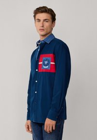 Camicia blu navy con colletto in denim, toppa rossa con strisce bianche e un design in blu sull taschino. Materiale in cotone.