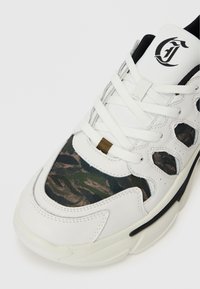 Weiße Sneaker mit Camouflage-Stoffpaneelen, weißen Schnürsenkeln, schwarzem Innenfutter und schwarzem Logo auf der Zunge.