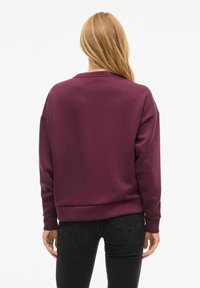 Donna bionda che indossa una felpa a maniche lunghe di un viola scuro e jeans neri, vista di spalle su uno sfondo bianco uniforme.