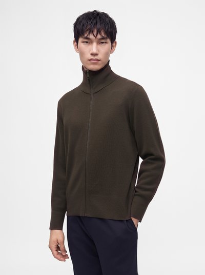 Jeune homme aux cheveux courts et foncés portant un pull à col montant zippé vert olive et un pantalon foncé, debout devant un fond clair uni.