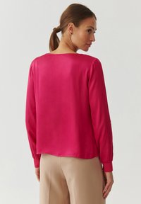 Blusa de manga larga fucsia con cuello redondo y textura satinada suave. Presenta un corte relajado y mangas con puños. Se combina con pantalones beige.