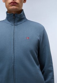 Blauwe zip-up jas met een hoge kraag, textuurstof en een klein geborduurd logo met een vlag op de linkerborst.