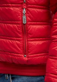 Röd isolerad jacka med horisontell quiltad design, silverfärgad dragkedja och ribbad midja. Har metalllogotypdetalj på dragkedjans draghandtag.