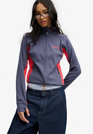 Ung kvinde med mørkt hår iført briller, en blå og rød lynlåsjakke og mørkeblå vidde jeans mod en hvid baggrund.