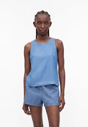 Femme aux cheveux tressés portant un haut bleu sans manches et un short bleu à fines rayures sur un fond blanc uni.