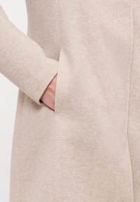 Beige strukturierter Mantel mit glatter Oberfläche, ausgestattet mit einerseitlichen Tasche und einem figurbetonten Design, das die Arm- und Ärmeldetails betont.