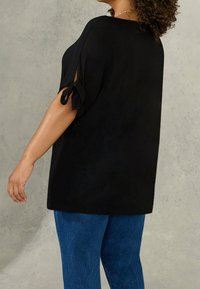 Black top with short sleeves featuring tied accents at the cuffs, paired with blue jeans. The fabric appears soft with a relaxed fit.