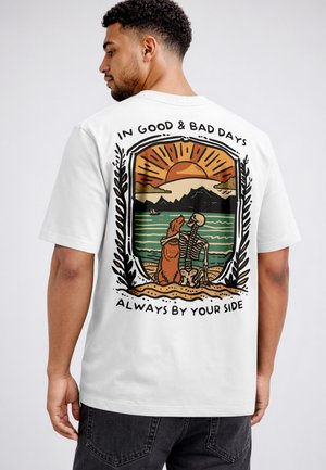 Man wearing white t-shirt with colorful graphic of a skeleton and dog sitting by a lake at sunset, text reads "In good & bad days always by your side."