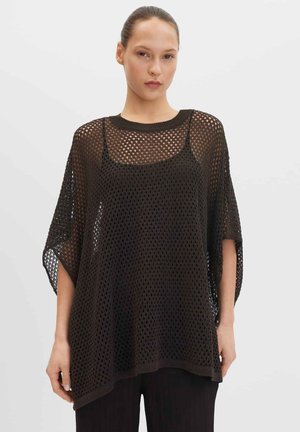 Junge Frau mit zurückgebundenem Haar, die ein lockeres, schwarzes, kurzärmeliges, offen gestricktes Oberteil über einem enganliegenden schwarzen Tanktop und schwarzen Hosen trägt.