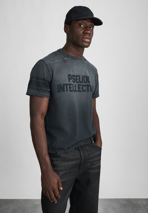 Black cotton t-shirt with "PSEUDO INTELLECTUAL" print, crew neck, short sleeves, distressed details. Paired with dark denim jeans.