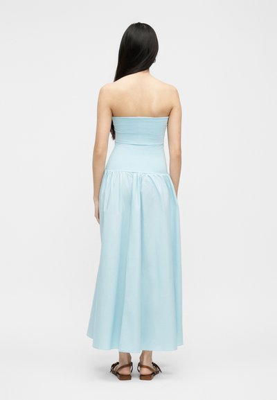 Γυναίκα με μακριά σκούρα μαλλιά που φοράει ανοιχτό μπλε μάξι φόρεμα χωρίς τιράντες και καφέ επίπεδα σανδάλια, στέκεται με την πλάτη γυρισμένη σε λευκό φόντο.