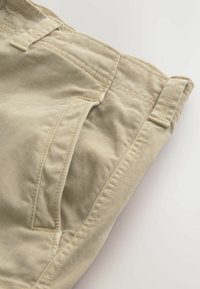 Détail d'un pantalon en coton beige montrant la poche avant, la passants de ceinture et les coutures sur un fond clair.