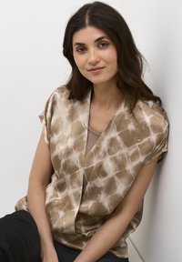 Blusa marrone a fantasia con una texture morbida e satinata, caratterizzata da un design ampio e maniche corte. Indossata sopra un top beige.