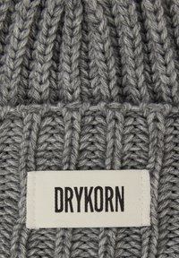 Bonnet gris en tricot présentant une texture côtelée et une étiquette blanche avec le nom de la marque "DRYKORN" en lettres noires en gras à l'avant.