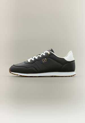 Baskets basses noires avec lacets blancs, languette de talon blanche, semelle en gomme et logo "GAP" doré sur le côté, sur un fond uni.