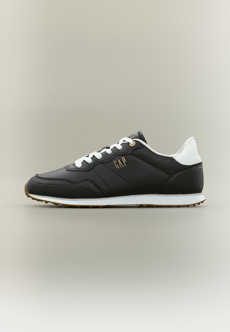 Baskets basses noires avec lacets blancs, languette de talon blanche, semelle en gomme et logo "GAP" doré sur le côté, sur un fond uni.