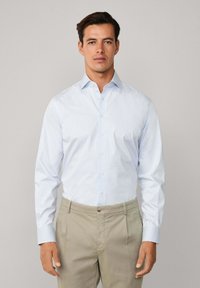 Muškarac u svijetloplavoj kariranoj košulji i bež hlačama, stoji nasuprot jednostavne svijetlo sive pozadine, okrenut prema naprijed.