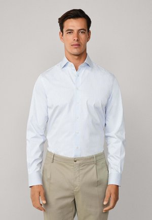 Mann in einem hellblauen karierten Hemd und beigen Hosen, stehend vor einem einfarbigen hellgrauen Hintergrund, frontal blickend.