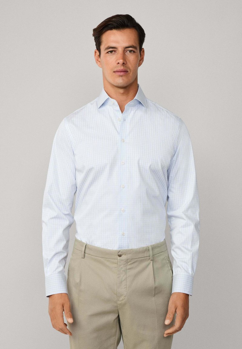 Muškarac u svijetloplavoj kariranoj košulji i bež hlačama, stoji nasuprot jednostavne svijetlo sive pozadine, okrenut prema naprijed.