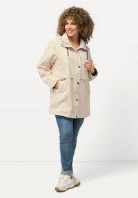 Giacca leggera beige con cappuccio, dotata di tasche laterali, chiusure a scatto e una vestibilità rilassata. Abbinata a jeans blu e sneakers bianche.