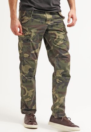 Man draagt groene en bruine camouflage cargobroek en bruine sneakers en staat voor een witte achtergrond.