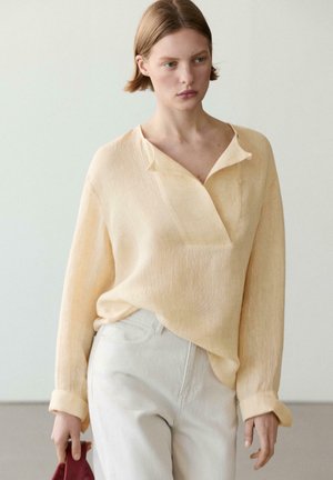 Young woman wearing a loose pale yellow blouse tucked into high-waisted white pants, holding a folded red fabric in her left hand.