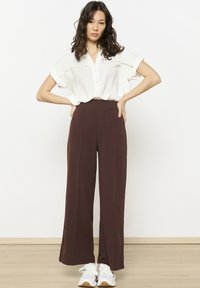 White short-sleeve button-up shirt with lace accents, paired with brown wide-leg pants and white sneakers. Wood floor background.