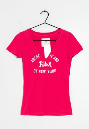 Kirkkaan pinkki, lyhythihainen V-kaula-aukkoinen T-paita, jossa teksti "Abercrombie and Fitch of New York", ripustettuna mustalle henkareille valkoista taustaa vasten.