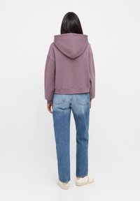 Korte paarse hoodie met een grote capuchon, ribbelrand en ruime mouwen, gecombineerd met hoge lichtblauwe jeans en beige sneakers.
