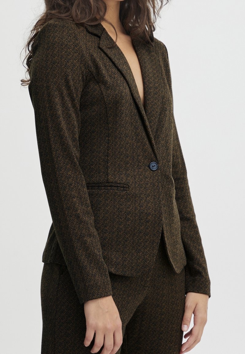 Femme portant un blazer marron à chevrons avec un seul bouton noir, assorti à un pantalon coordonné, sur un fond clair uni.