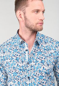 Camicia a maniche corte blu con fiori, caratterizzata da uno sfondo blu navy con motivi bianchi e azzurri chiari, colletto a punta e bottoni scuri.