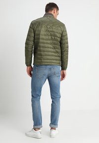 Olivengrøn quilted pufferjakke med ståkrave, ribbede detaljer og en tætsiddende pasform. Båret med lyseblå jeans og hvide sneakers.