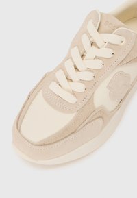 Beige mocka och krämfärgad läder sneakers med platta snören, vadderad tunga och präglad logodetalj på sidan. Litet rundad tådesign.