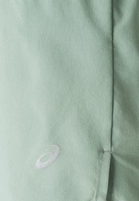 Light green athletic shorts made of smooth fabric featuring a silver logo and stitched hem detail. Shows texture and color clearly.