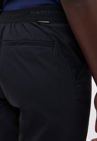 Gros plan sur une personne portant un pantalon bleu marine avec une poche passepoilée arrière et une ceinture noire portant l'inscription "Peak Performance".