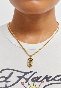 Gullkjede med et rektangulært anheng med et detaljerte design. Båret over en lysfarget, trykket t-skjorte.