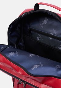 L'intérieur de ce sac synthétique rouge est doté d'une doublure sombre avec un motif de logo répétitif, d'une poche zippée et d'un compartiment principal spacieux.