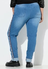 Jeans de mezclilla azul claro de corte ajustado, con una cinta de logo en blanco y negro a lo largo de los lados y con los puños doblados. Dos bolsillos traseros visibles.