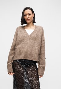 Καφέ πουλόβερ πλεκτό σε υπερμεγέθη γραμμή με λαιμόκοψη σε σχήμα V, ριμπ τελείωμα και λεπτομέρειες με κουμπιά στα μανίκια, φορεμένο πάνω από μια μαύρη φούστα με παγιέτες.