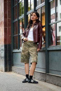 Ung kvinde i en bordeaux læderjakke, hvid top, camouflage shorts og sorte sneakers som læner sig op ad en bygning med glasvinduer.