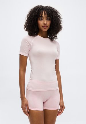 Jeune femme aux cheveux bouclés portant un haut ajusté à manches courtes rose clair et un short assorti, debout devant un fond blanc uni.