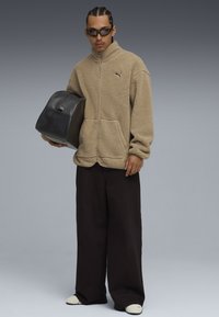 Chaqueta de forro polar con cremallera en color beige, con cremallera frontal y bolsillos laterales. Bolsa de cuero negro en una mano, combinada con pantalones negros holgados y zapatos blancos.