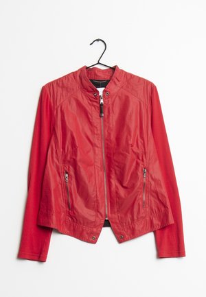 Veste rouge pour femme avec fermeture éclair intégrale à l'avant, col montant, poches latérales zippées et manches longues, suspendue à un cintre noir sur fond blanc.