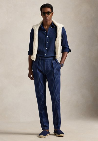 Hombre con camisa azul oscuro con botones, pantalones azules a rayas finas, suéter blanco sobre los hombros, gafas de sol y alpargatas azules.