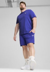 Královsky modré sportovní tričko a šortky vyrobené z měkké tkaniny, doplněné o bílé tenisky a bílé ponožky po kotníky. Zobrazené logo značky.