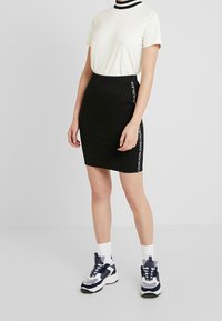 Svart kjol med vitt logodetalj längs sidan, ihop med en krämfärgad kortärmad topp med en svart randig krage. Sneakers avslutar looken.